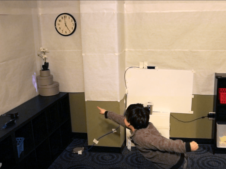 A child enthusiastically points at a projected digital character peeking out from behind some decorative boxes on a shelf.